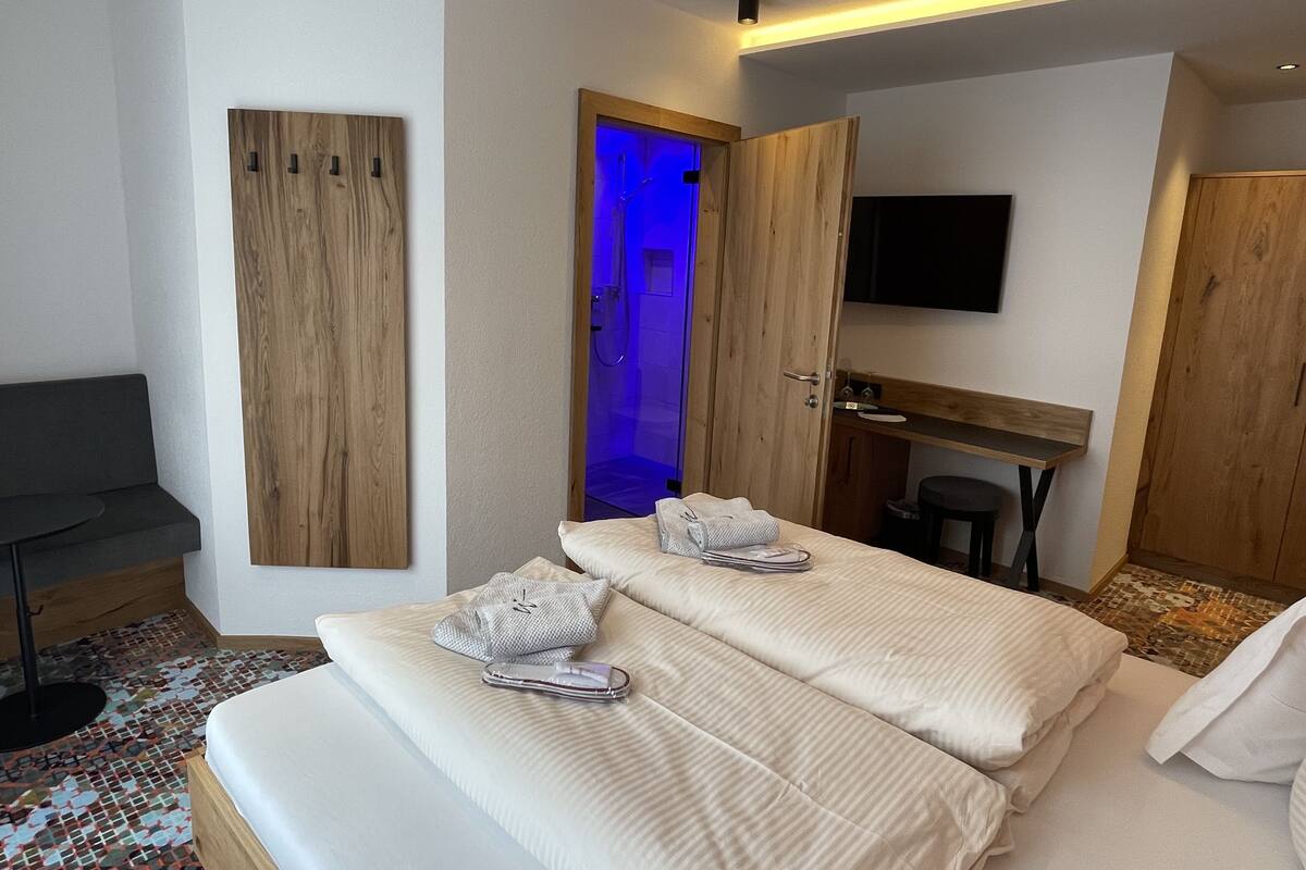 A hotel room with a bed, a desk, a chair, a TV, and a bathroom with a blue-lit shower.