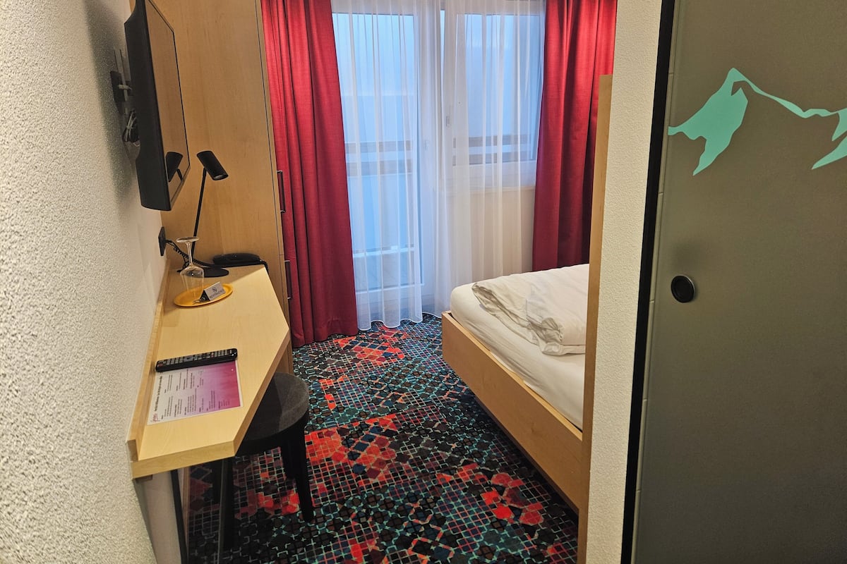 A compact hotel room with a bed, a desk with a chair, a lamp, and a window with red curtains.