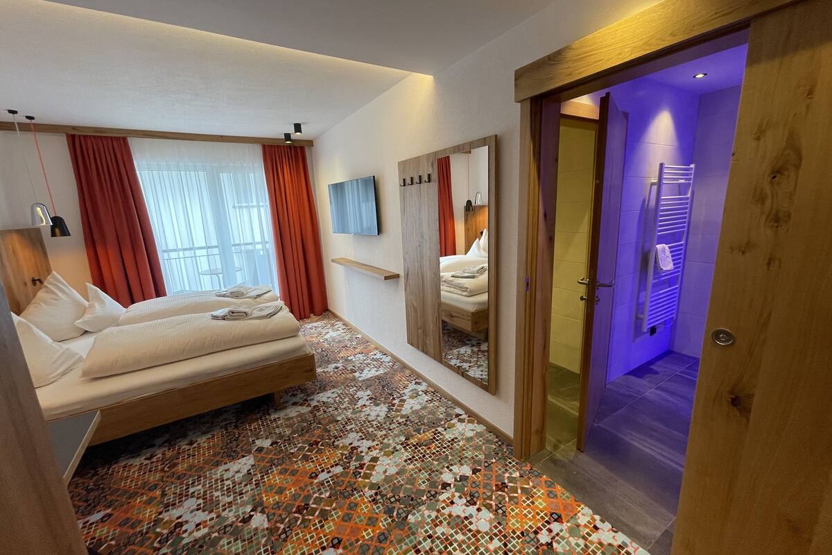 A hotel room with a bed, a mirror, a TV, and a bathroom with a shower and a towel rack.