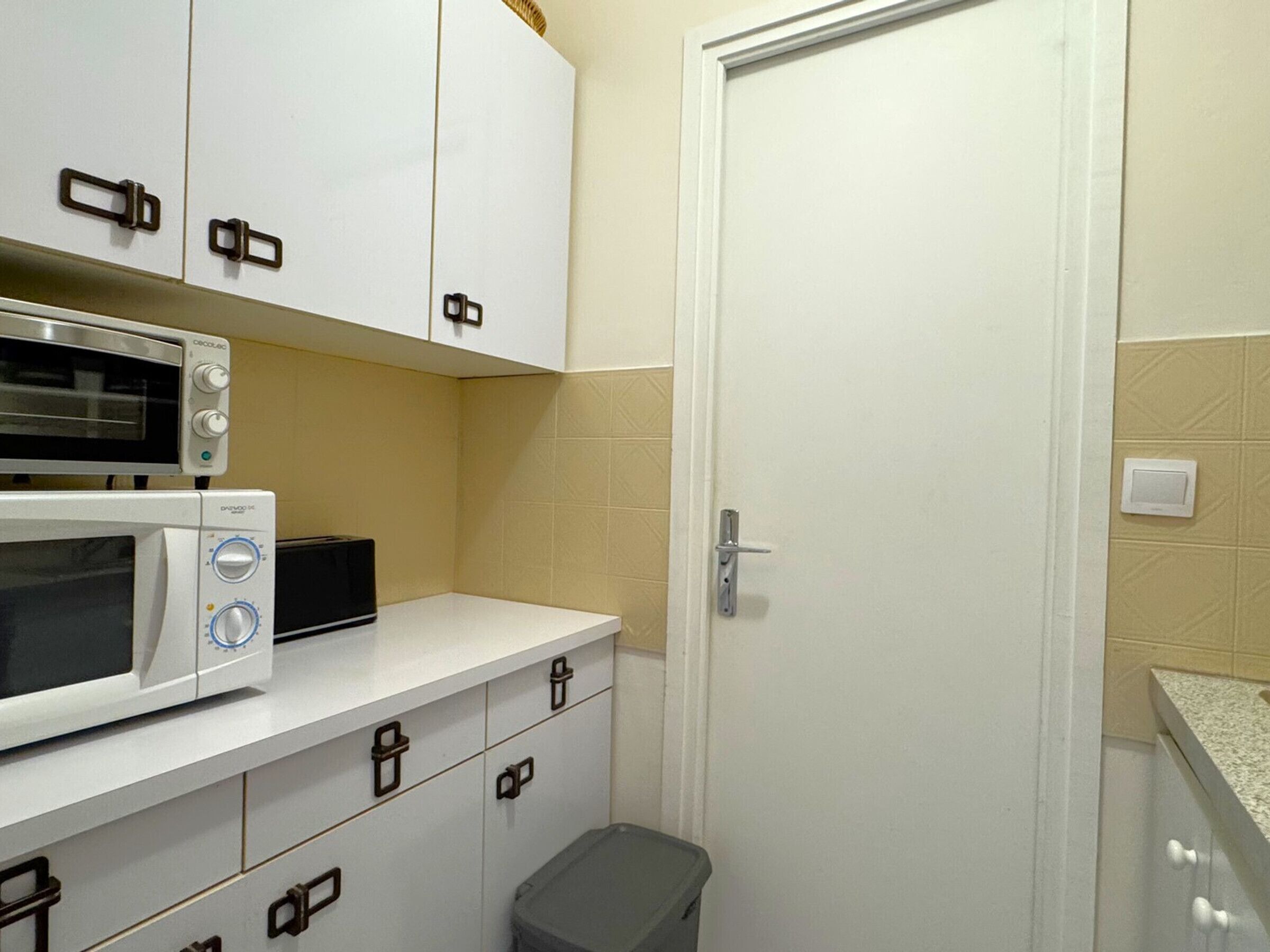 Una cocina compacta con armarios blancos, un microondas, un tostador y un cubo de basura.