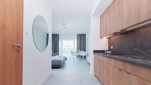 Una habitación de hotel moderna con puerta de madera, un espejo redondo, una kitchenette con fregadero y estufa, un comedor con mesa y sillas, y una cama con cubrecama gris.