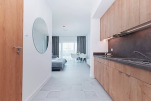 Une chambre d’hôtel moderne avec une porte en bois, un miroir rond, une kitchenette équipée d’un évier et d’une cuisinière, un coin repas avec une table et des chaises, et un lit recouvert d’une couverture grise.