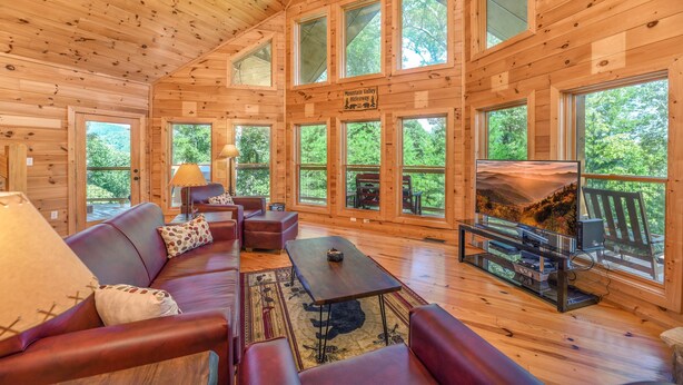 A cozy cabin interior with a wooden interior, large windows, a sofa, a table, and a fireplace. - 🏔️ Mountain Valley Hideaway 🌳 Elevated Deck, 2 King Suites & Hot Tub (Sevierville)