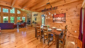 A wooden cabin interior with a dining table set for four, wooden walls, and large windows. - 🏔️ Mountain Valley Hideaway 🌳 Elevated Deck, 2 King Suites & Hot Tub (Sevierville)