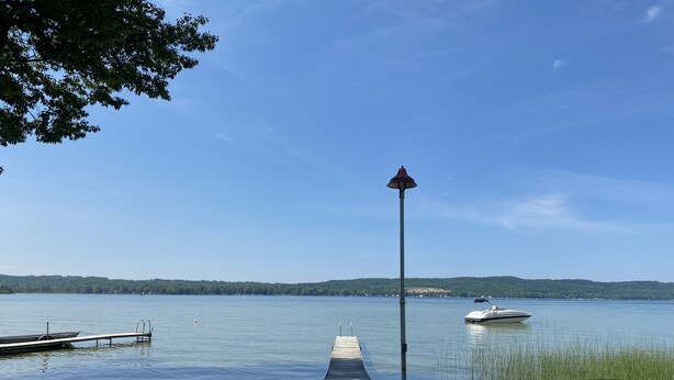 A lakeside dock with chairs and a bench, a boat on the water, and a distant shoreline.