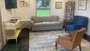 A living room with a grey sofa, a blue armchair, a wooden shelf with books, a small table, and a television mounted on the wall.