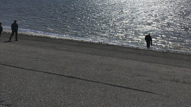 Una playa con cielo despejado, gente caminando por la orilla y cometas volando a lo lejos.
