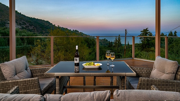 A covered outdoor dining area with wicker furniture, a table with a bottle of wine, glasses, and a plate of grapes. - Andromeda Luxury Villa (Zakynthos)