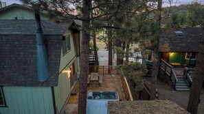 A rustic cabin with a wooden deck, a hot tub, and a view of a forested area. - Spacious Retreat with Hot Tub, Game Room, Meadow Views & 2 Fireplaces (Big Bear City)