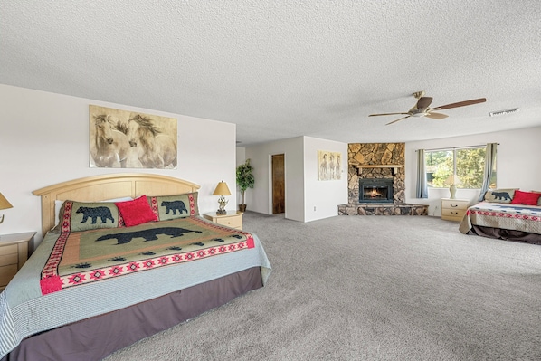 A spacious bedroom with a large bed, a fireplace, a ceiling fan, and a view of the outdoors.