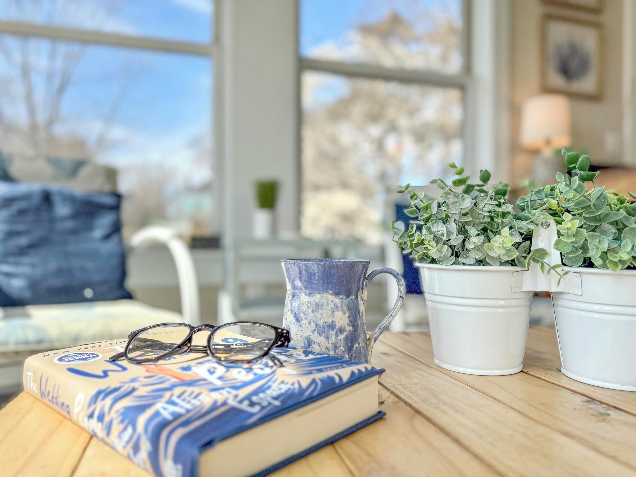 Une table en bois sur laquelle se trouvent un livre, une tasse bleue et blanche et deux plantes en pot blanches.