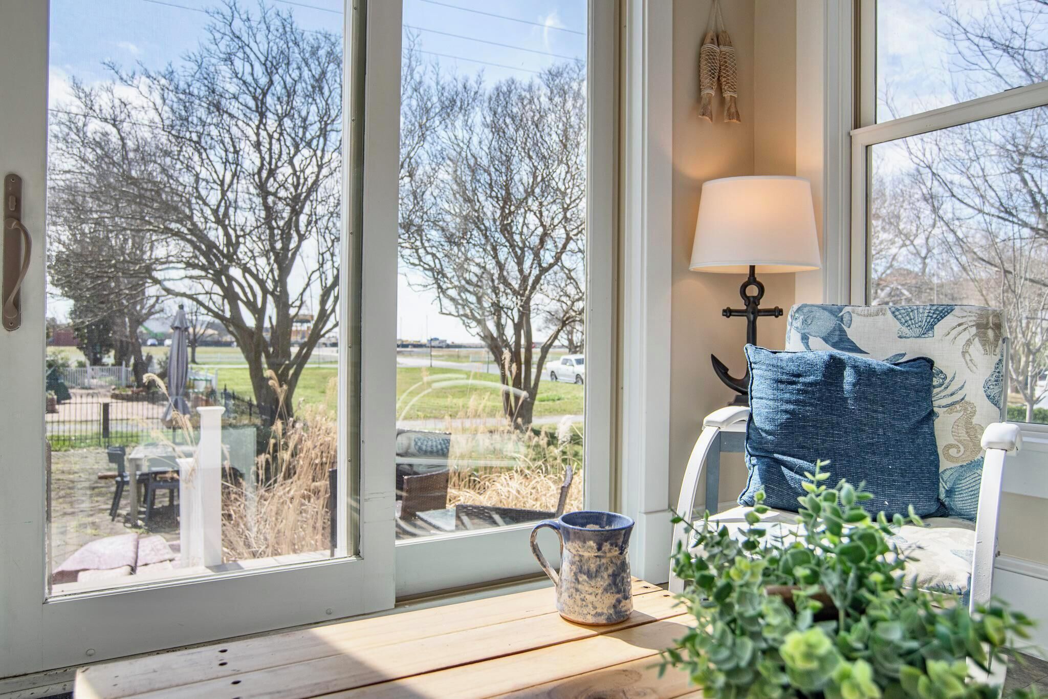 Ein Zimmer mit Blick durch ein großes Fenster, eine Lampe, ein Sessel mit Kissen und eine Pflanze auf einem Tisch.