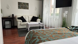 A hotel room with a bed, a sofa, a small table with a tablecloth, a TV mounted on the wall, and a window with sheer curtains. - L'Embleme Boutique Hotel (Beaurepaire)