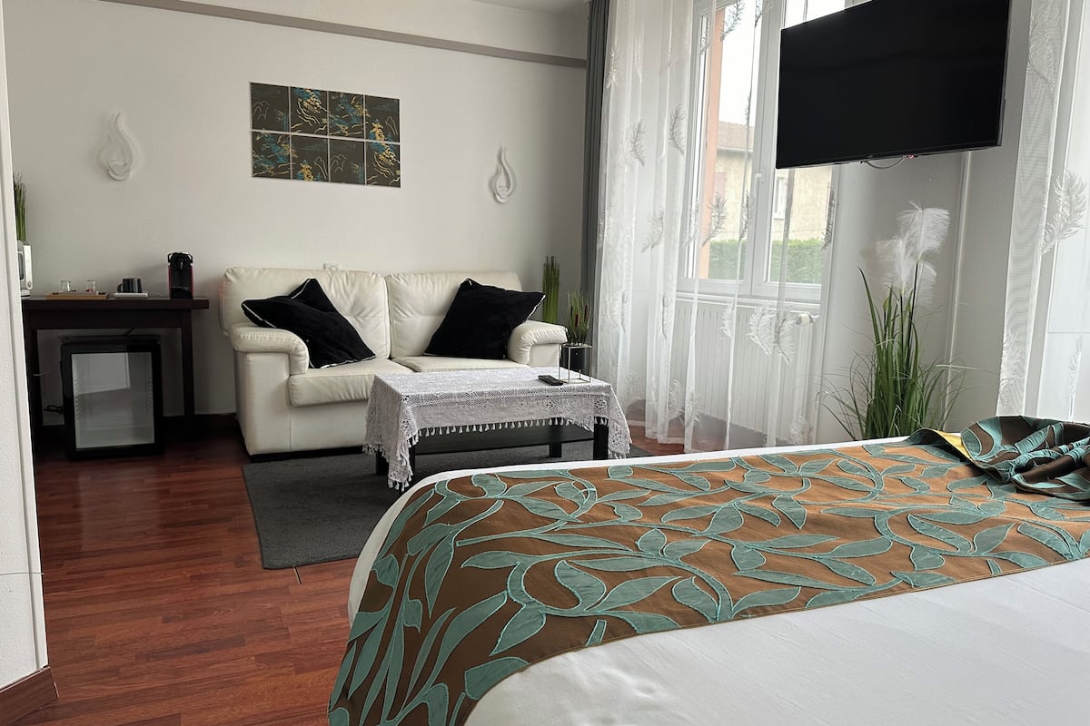 A hotel room with a bed, a sofa, a small table with a tablecloth, a TV mounted on the wall, and a window with sheer curtains.