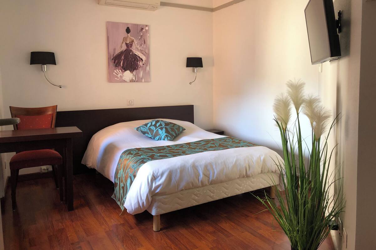 A hotel room with a bed, a desk, a chair, an air conditioner, a wall-mounted TV, and a plant.