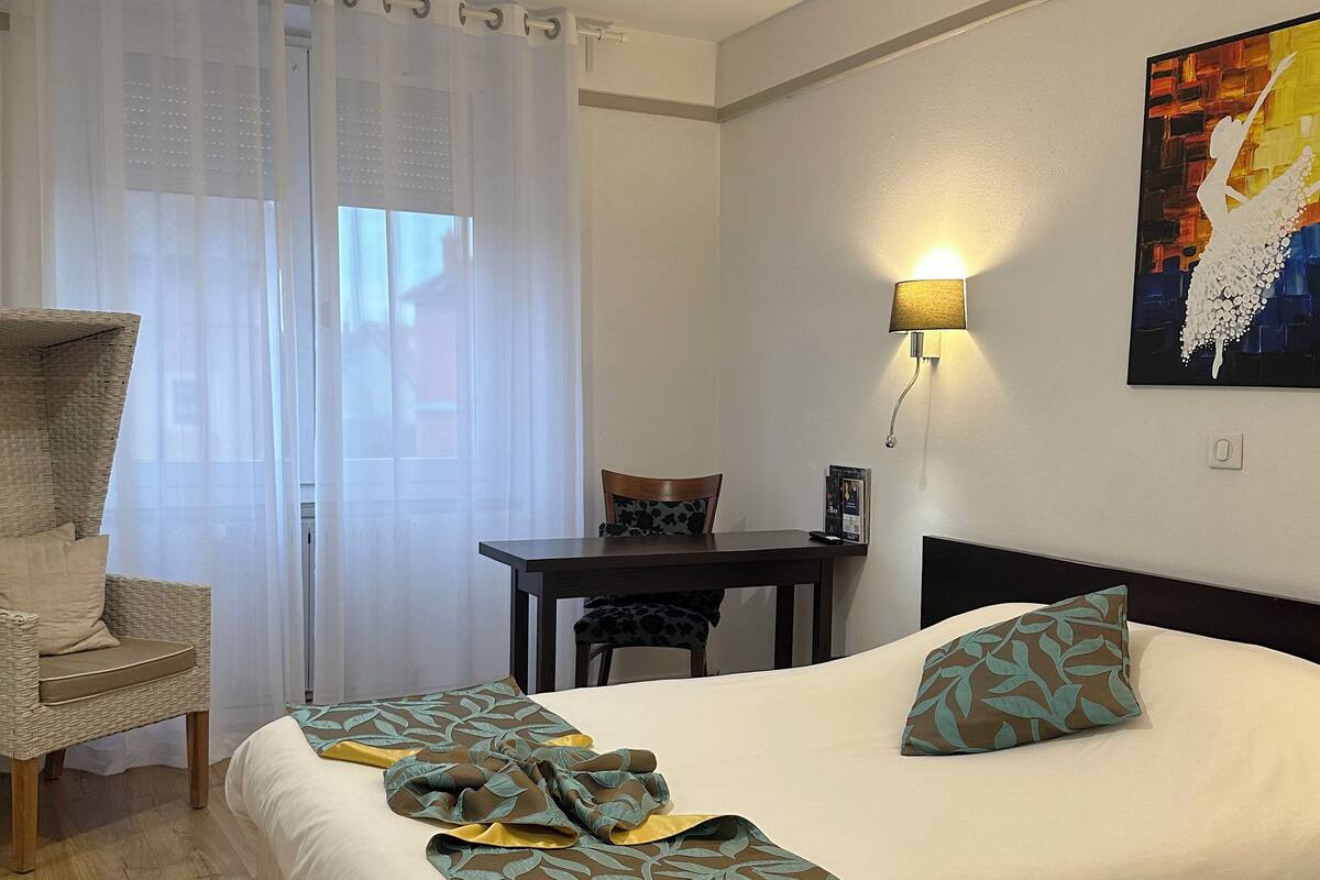 A hotel room with a bed, a chair, a table, and a painting on the wall.