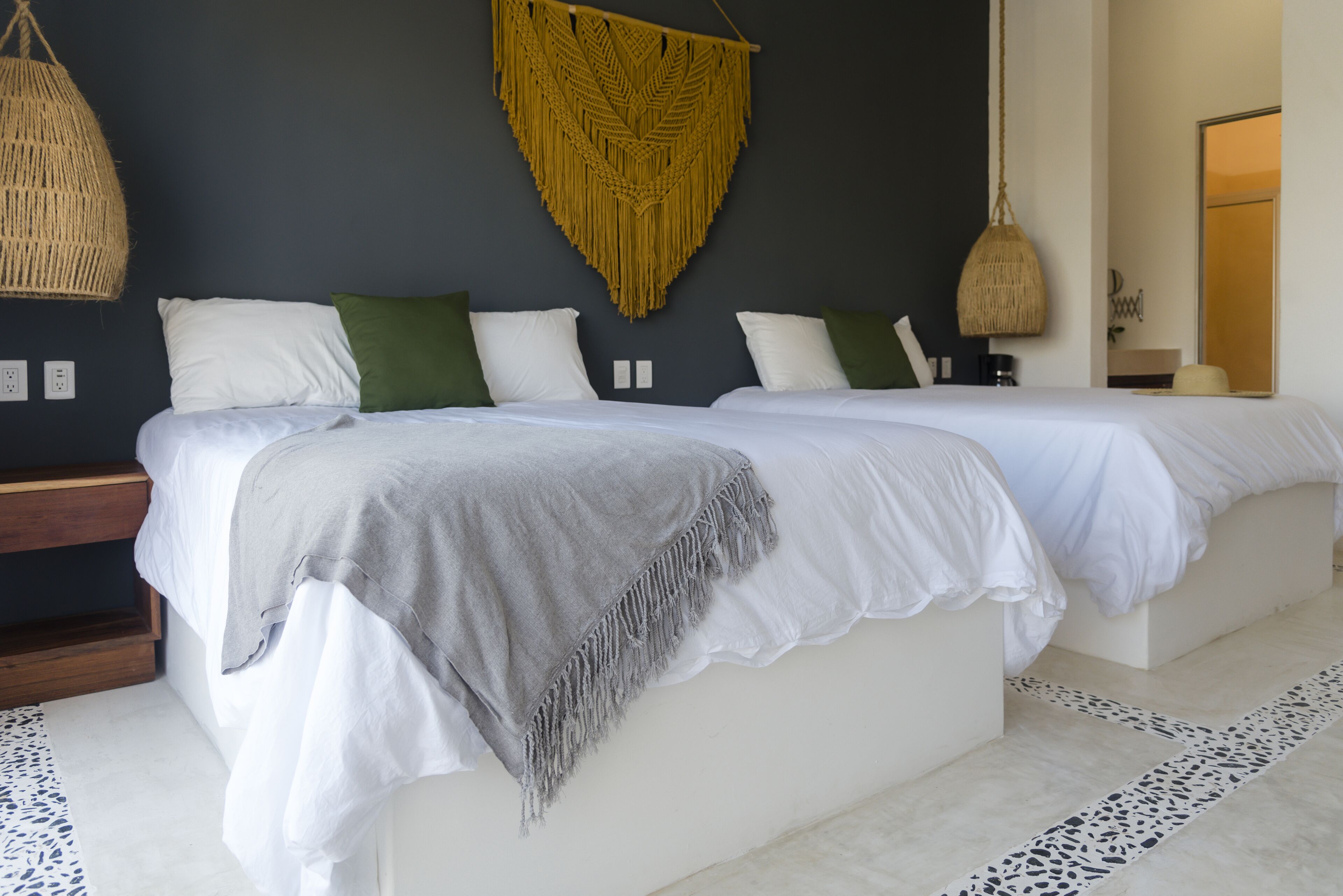 Una cama doble con ropa blanca y un plaid gris, un colgador de macramé y una lámpara colgante de tejido.