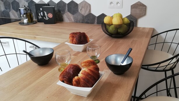 A wooden dining table with two bowls of croissants, two glasses of water, and a bowl with a spoon. A basket of lemons is on the table, and there are two chairs. - The Cabanon from the Pas du Loup (la seyne sur mer)