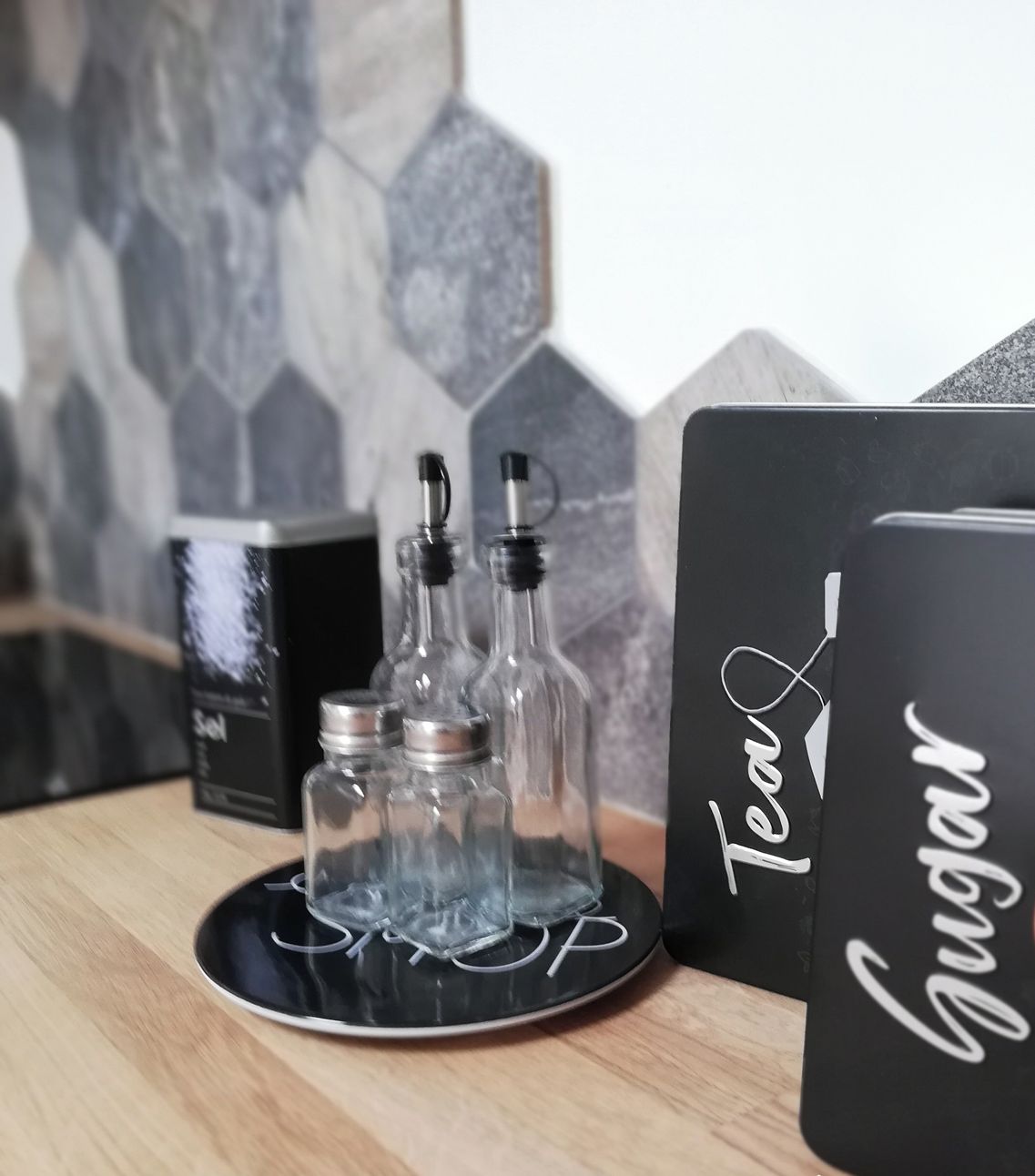 A kitchen counter with a black tray holding two glass bottles, a black box labeled "Tea," and a black box with "Gugar" and a sugar bowl illustration.