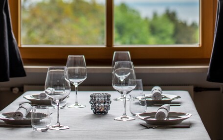A dining table set with wine glasses and plates, a small candle, and a window with a view of greenery and the sea.