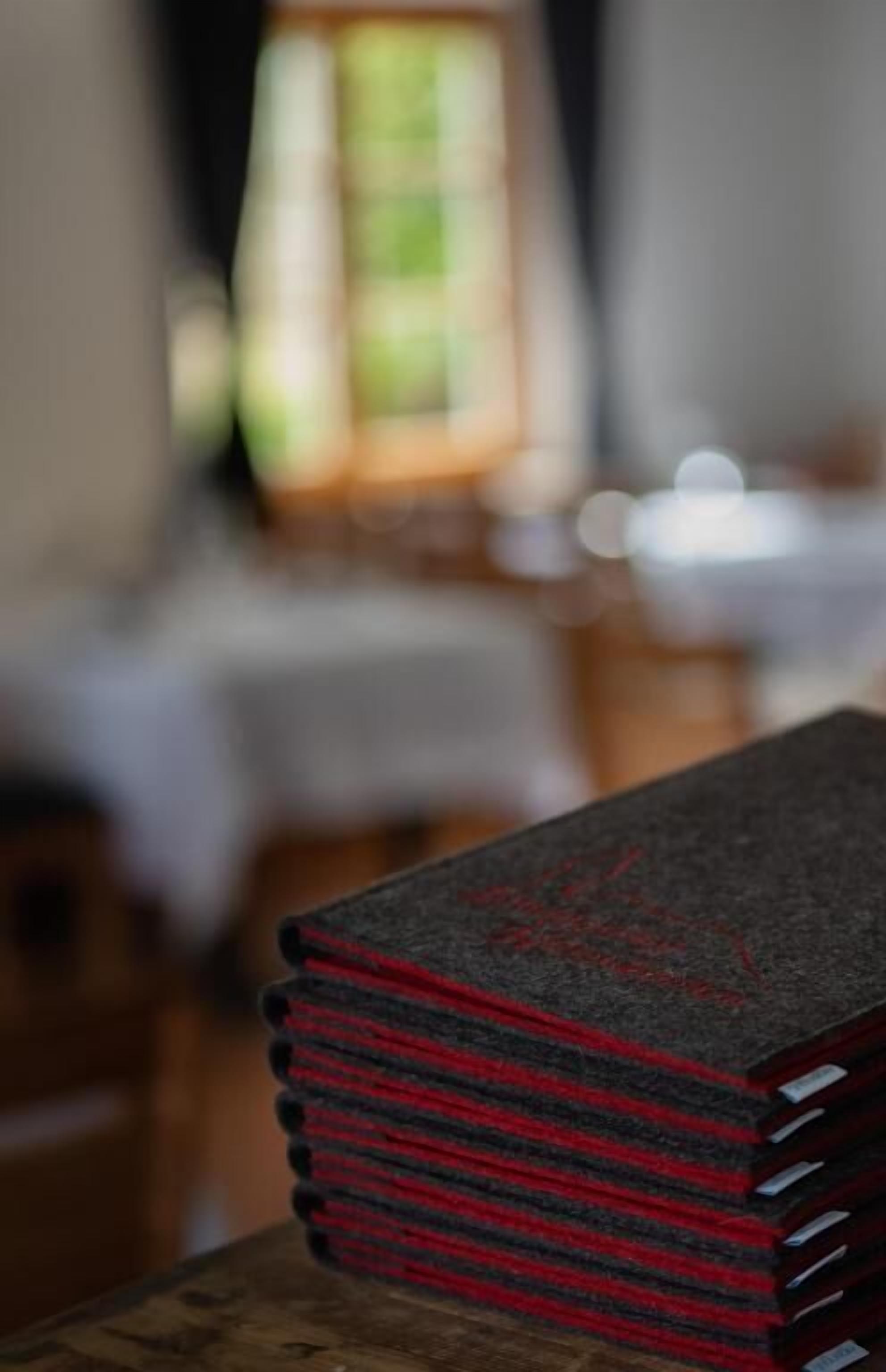 Una pila de llibres sobre una taula de fusta en una habitació amb una finestra i un fons desenfocat.