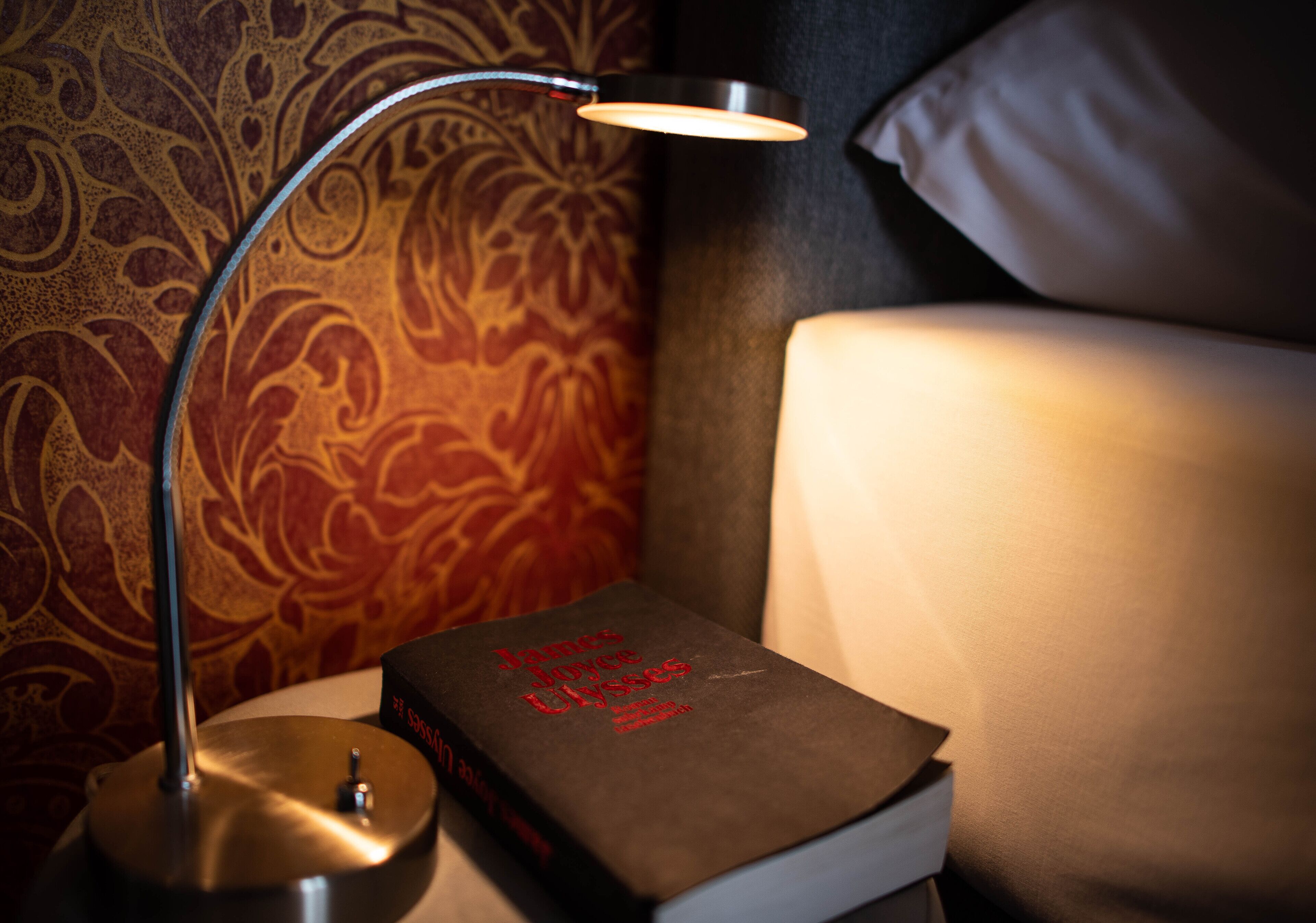 A bedside table with a lamp, a book titled "James Joyce Ulysses," and a patterned wall.