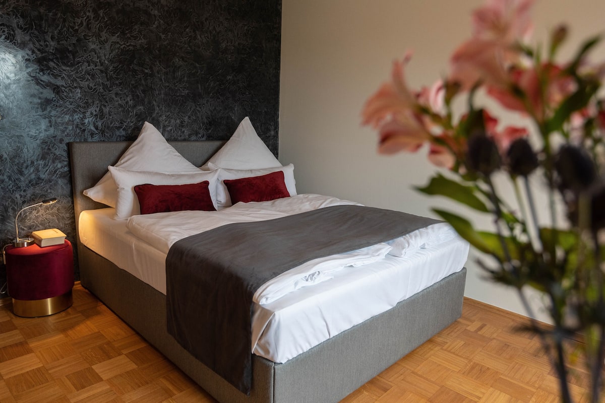 A modern bedroom with a grey upholstered bed, white linens, and red pillows. A small red ottoman and a bedside table with a lamp are visible.