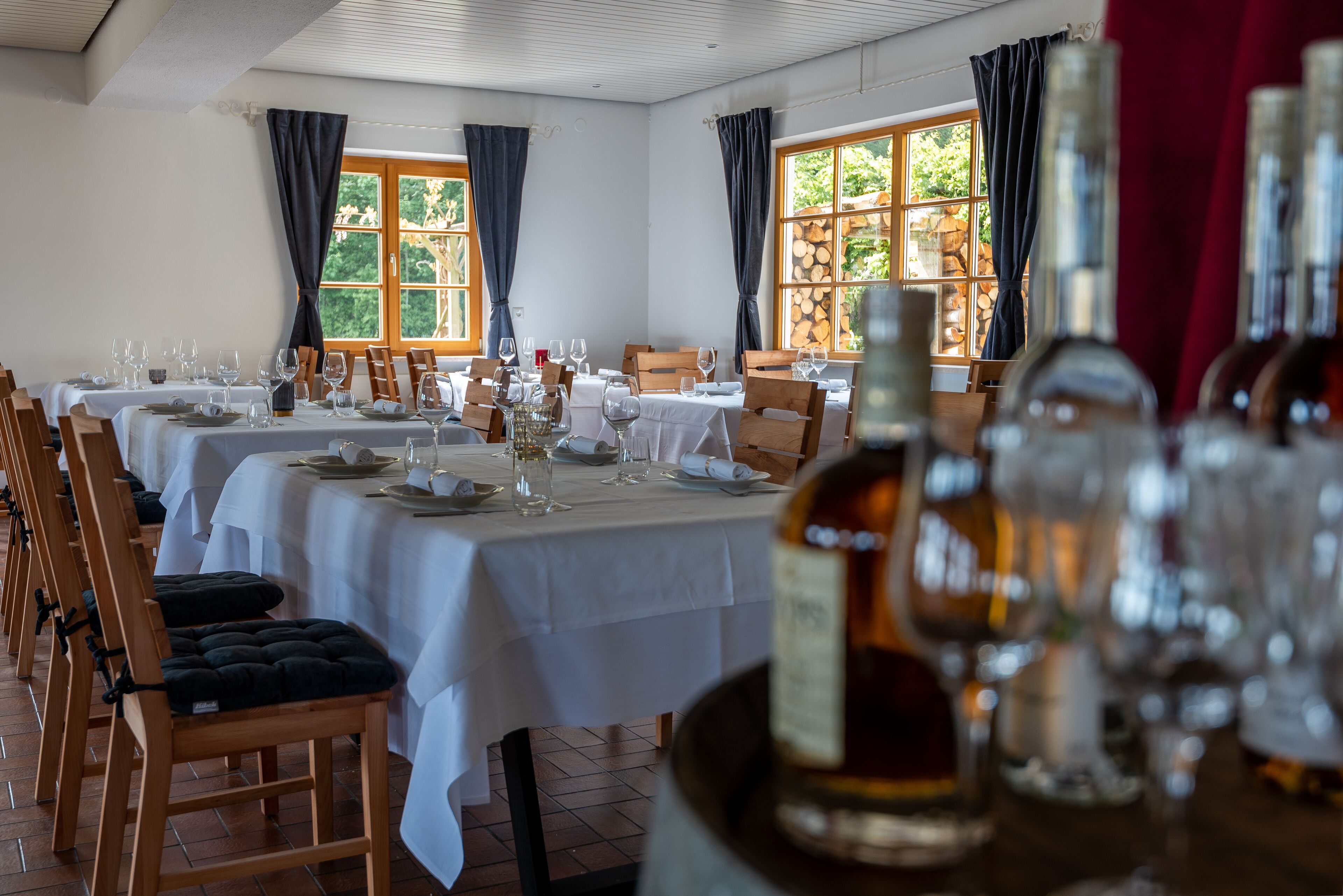 Un coin repas avec des tables dressées pour le repas, des chaises en bois et une bouteille de vin au premier plan.