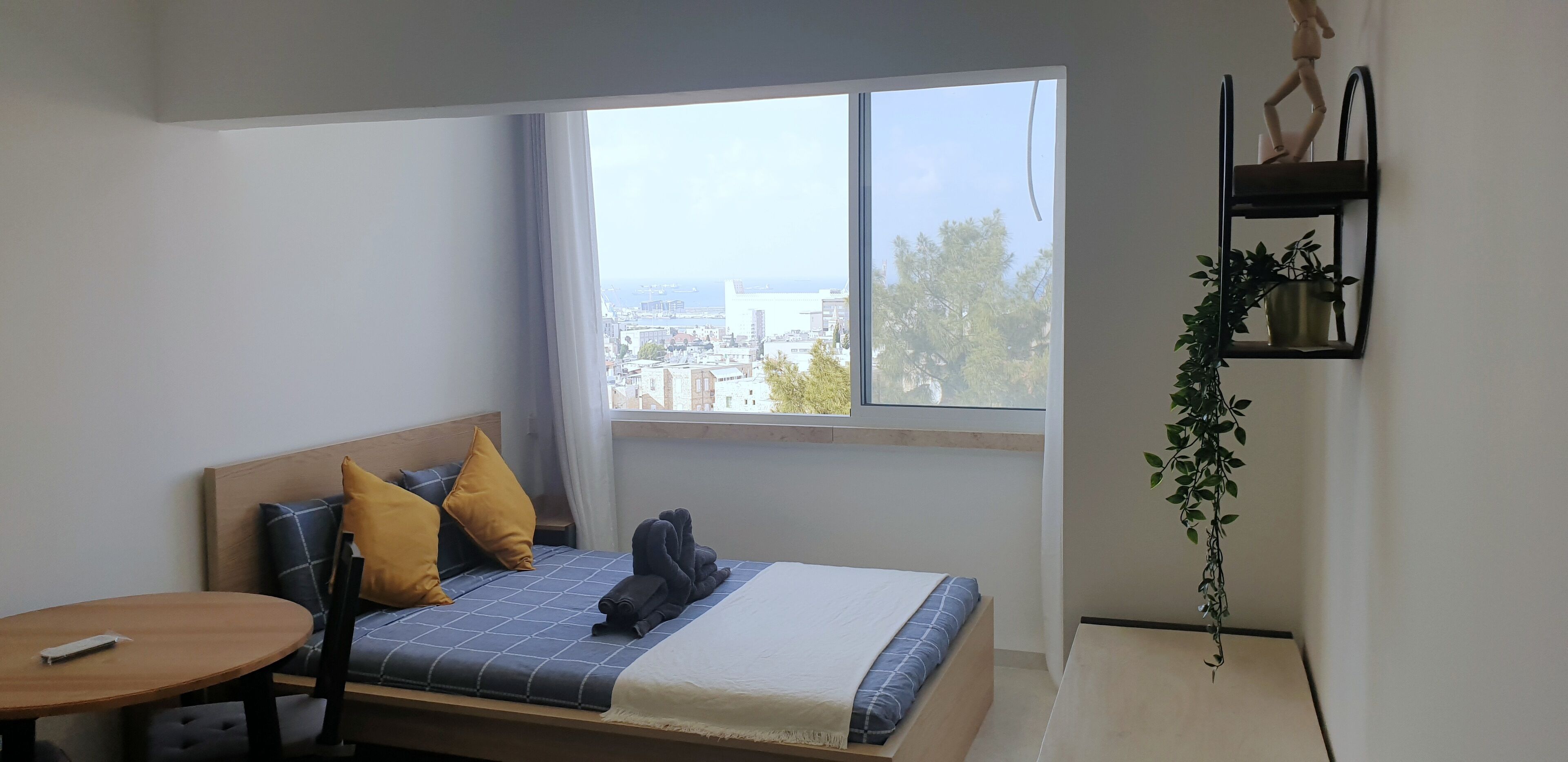 Una habitación de hotel moderna con un ventanal grande, una cama con ropa de cama azul, un cabecero de madera y una mesita.