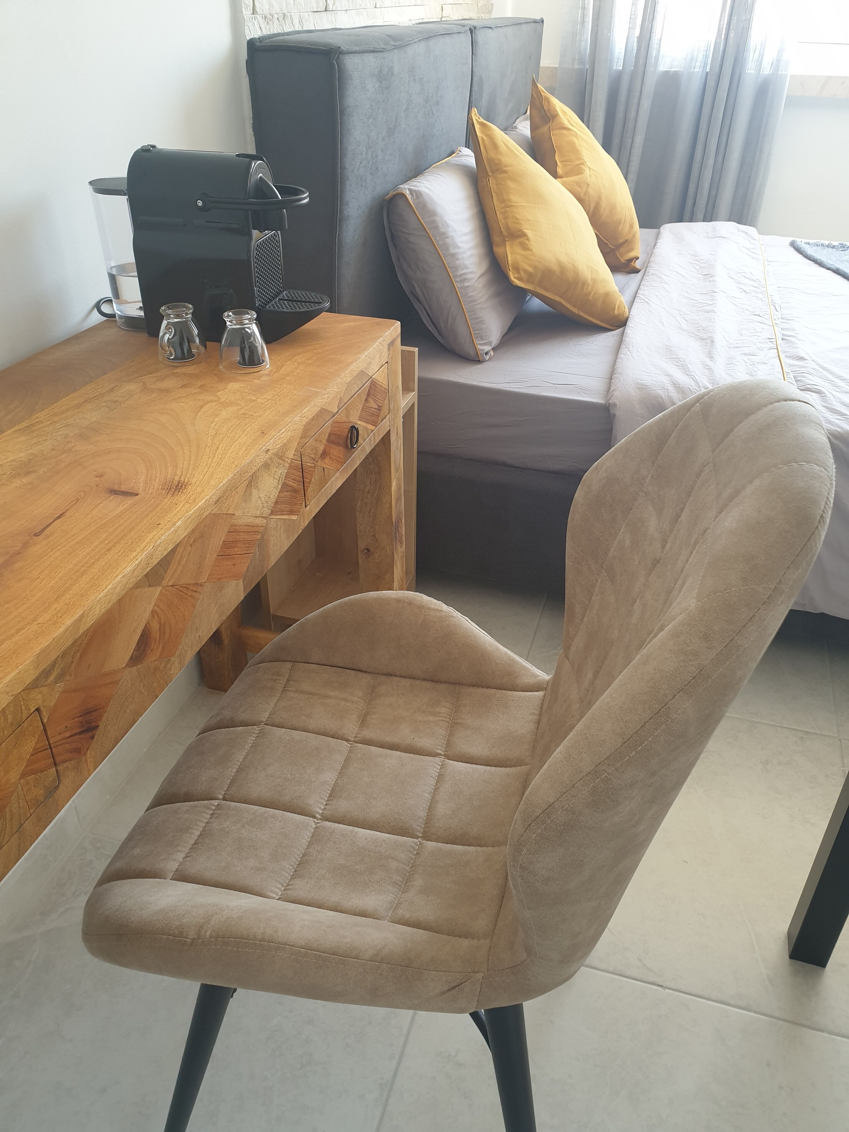 Um quarto moderno com escrivaninha de madeira, poltrona confortável, sofá cinza com almofadas amarelas e cama com edredom branco.