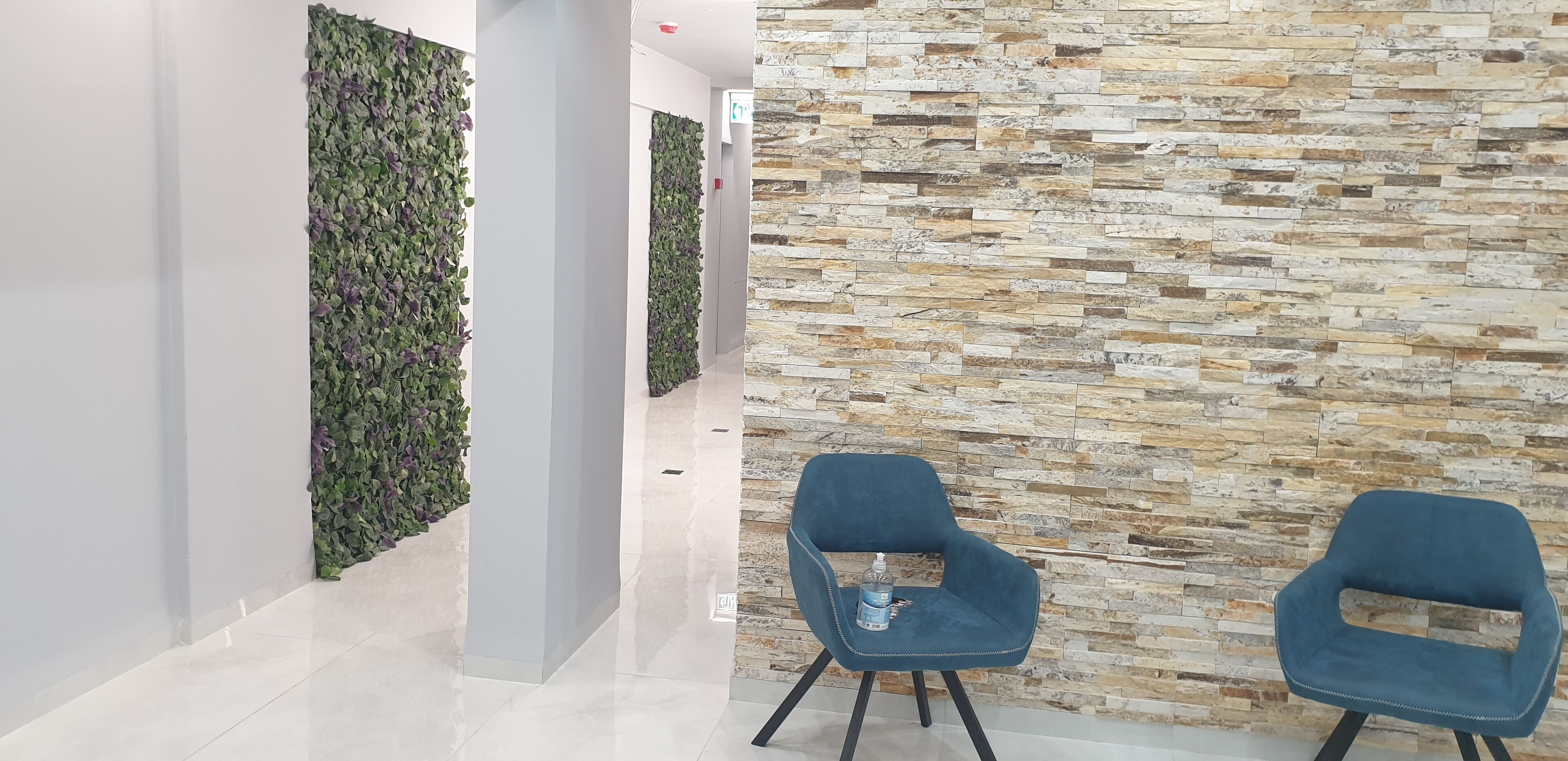 A hallway with a green leafy wall, a stone-textured wall, and two blue chairs with a water bottle on one.
