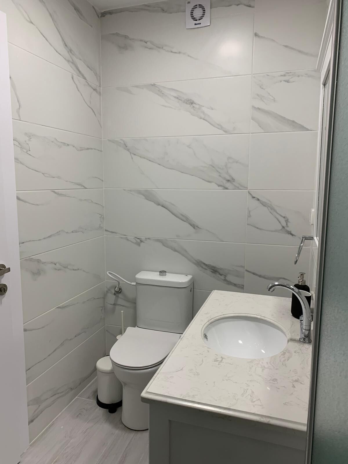 A bathroom with white marble tiles, a toilet, a sink with a mirror, and a white trash bin.