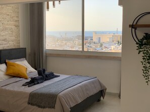 A bedroom with a large window, a bed with pillows and blankets, a headboard, and a hanging plant. - Maya Guest House Panoramic Sea&City view (Haifa)