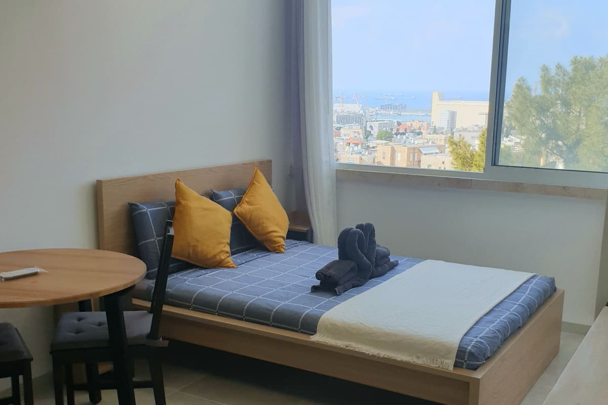 A single bed with a wooden frame, two pillows, a blanket, and a pair of folded towels. There is a small round table with two chairs, a window with a view of buildings and trees, and a curtain partially drawn.