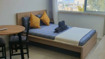 A single bed with a wooden frame, two pillows, a blanket, and a pair of folded towels. There is a small round table with two chairs, a window with a view of buildings and trees, and a curtain partially drawn.