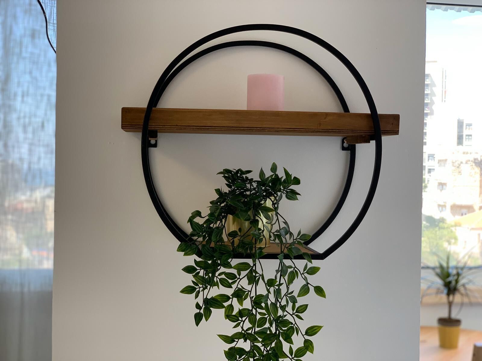 Un marco metálico circular montado en la pared con un estante de madera que sostiene una vela rosa y una planta en maceta.