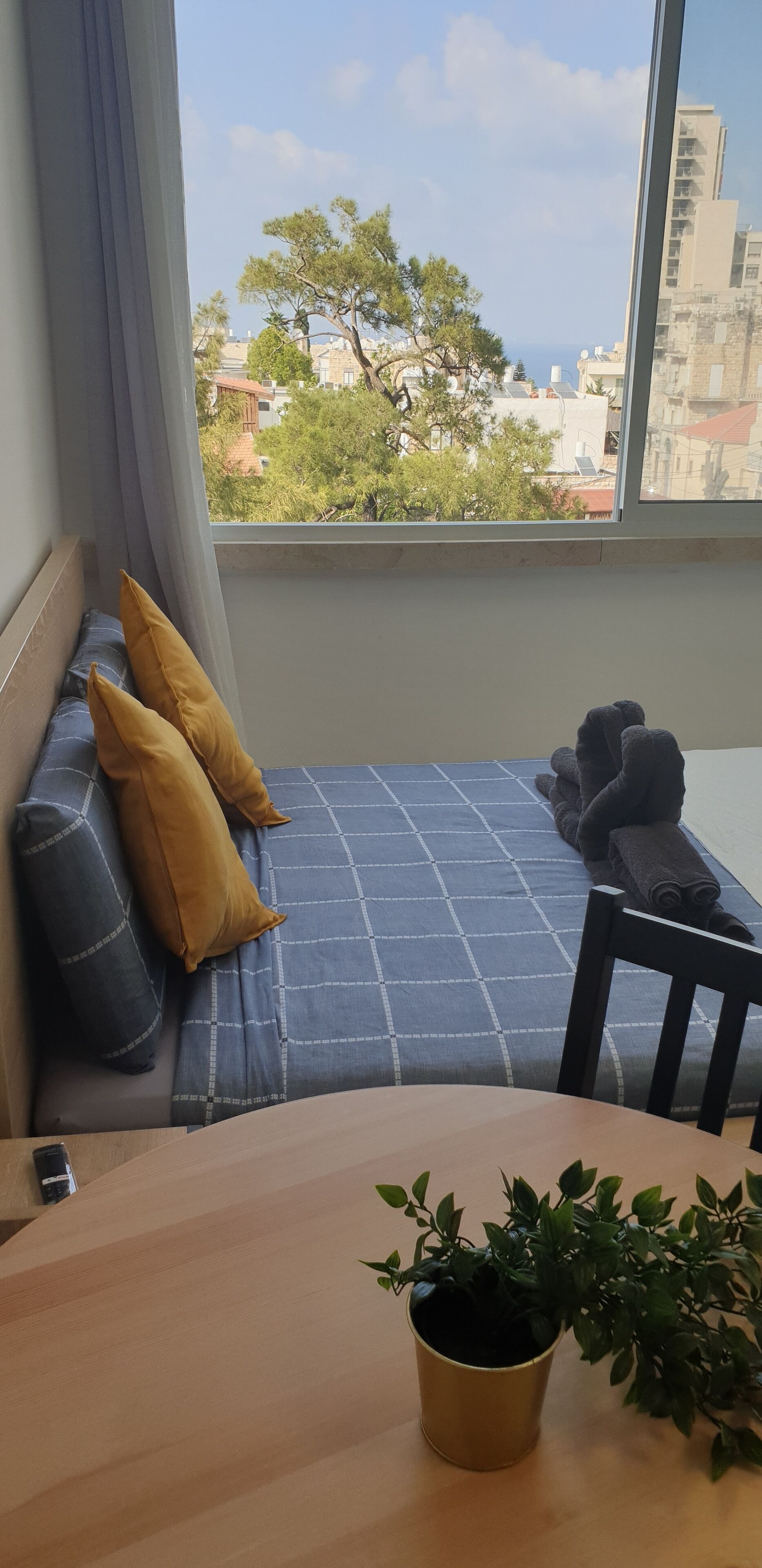 Una habitación con cama, una silla, una mesa y una ventana que da a vista de edificios y árboles.