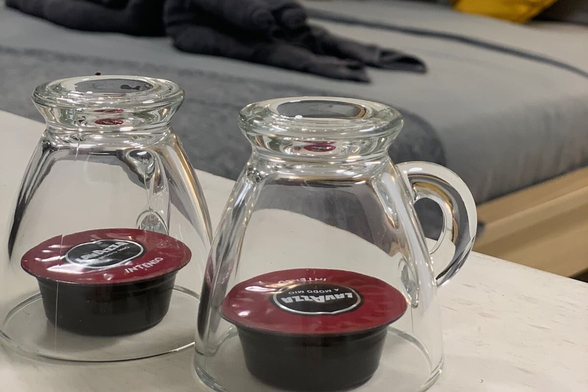 Two glass containers with red lids on a table in a room with a bed, pillows, and a stone wall.