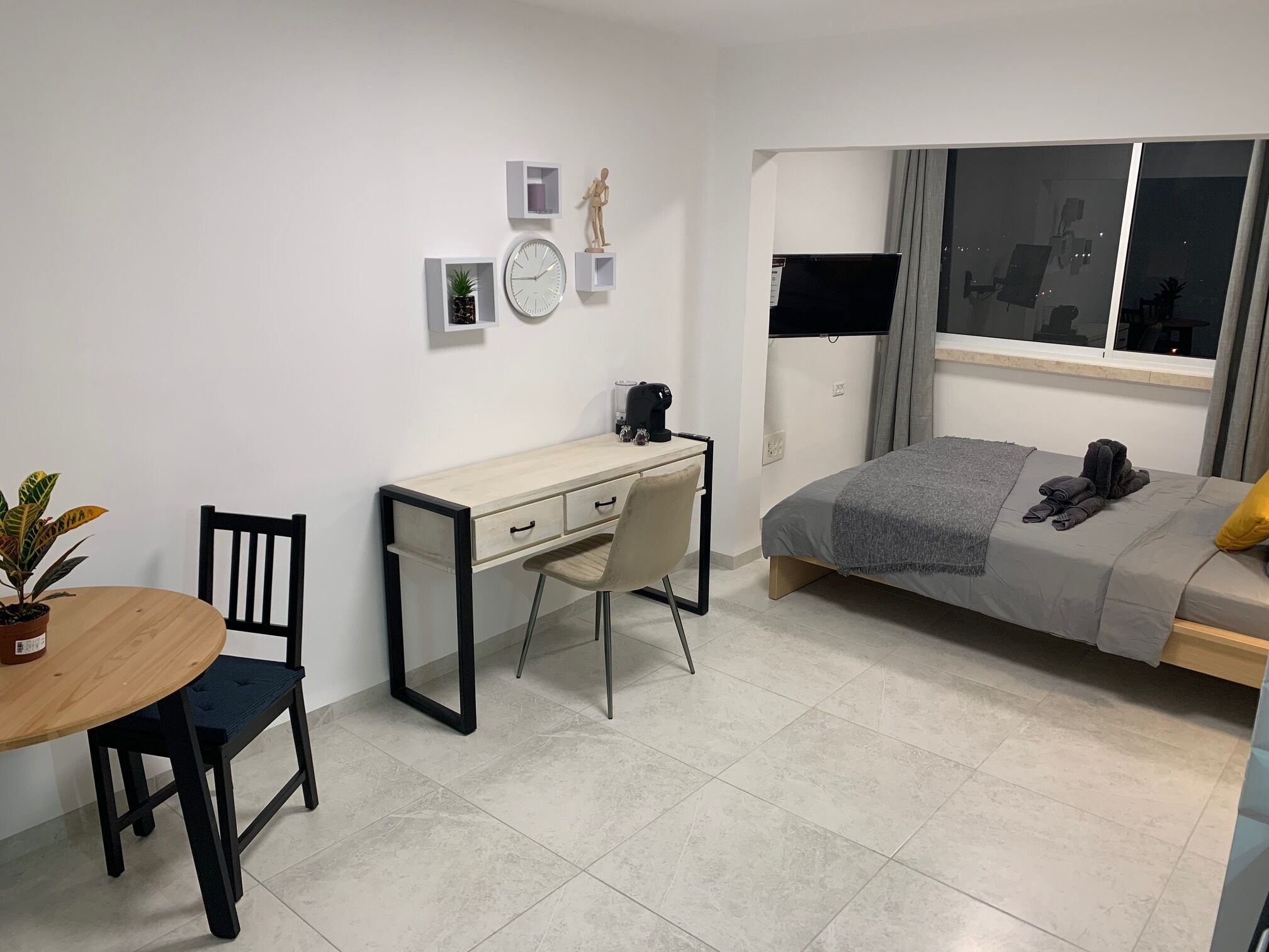 A modern bedroom with a bed, a desk, and a chair. There is a window with curtains, a potted plant, and a clock on the wall.