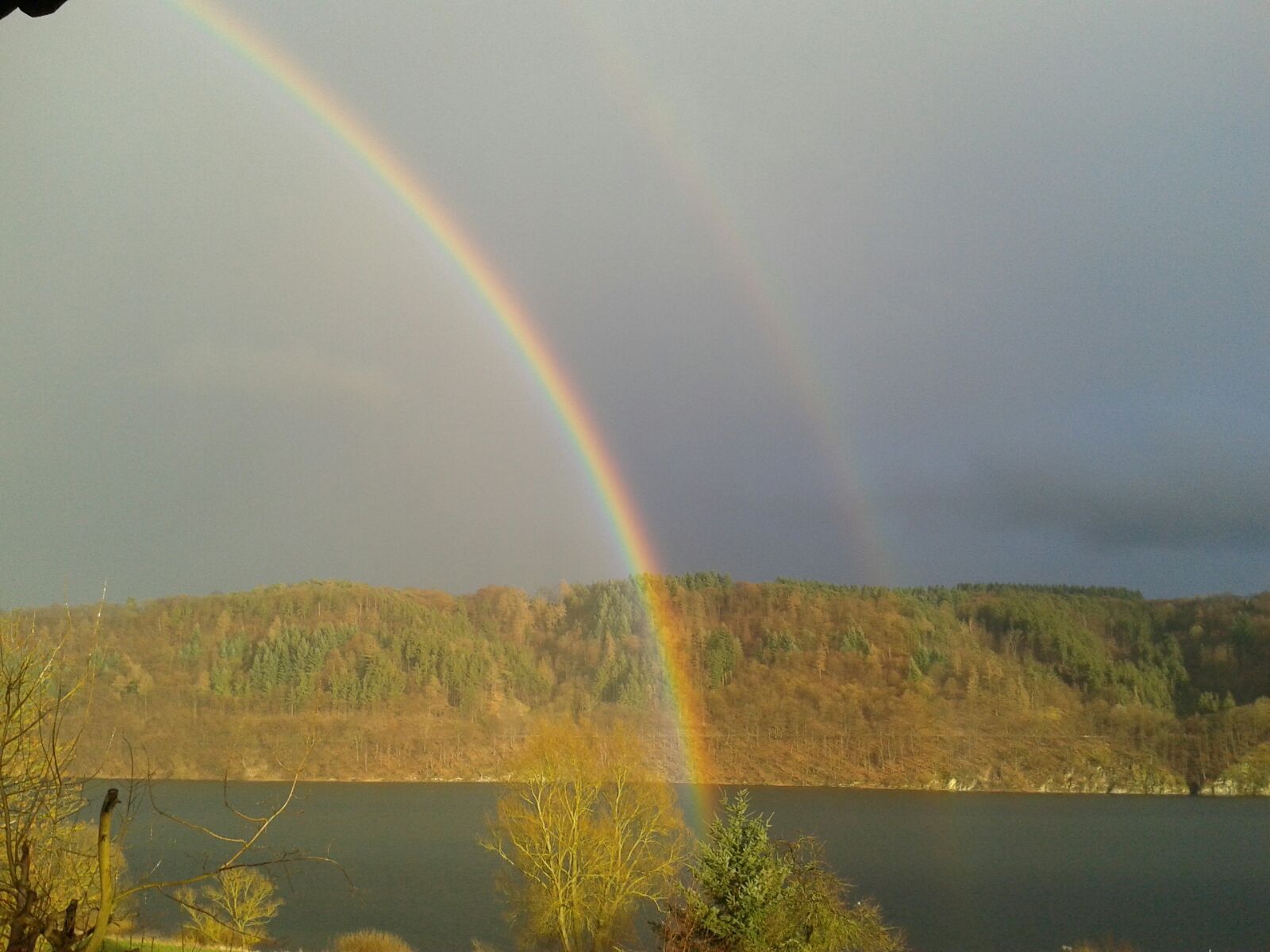A lake with a rainbow and a forested hill in the background.