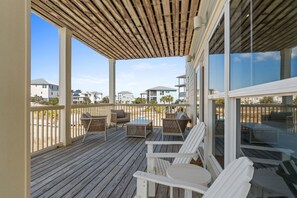 A covered outdoor deck with white Adirondack chairs, a table, and a glass railing overlooking a beach. - Dune Our Thing: Charming North Cape Home- Sleeps 14, New Private Pool! (Cape San Blas)