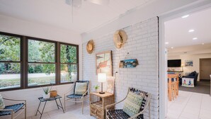 A room with a white brick wall, large windows, wicker furniture, and a television. - 1 Cassina-Lovely Beach Cottage, Steps to the Sand (Hilton Head Island)