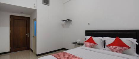 A hotel room with a bed, a wooden door, a small table, and a wall-mounted shelf.