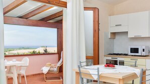Una cocina con armarios blancos y un microondas, un comedor con una mesa y sillas, y vista al mar a través de una puerta abierta.