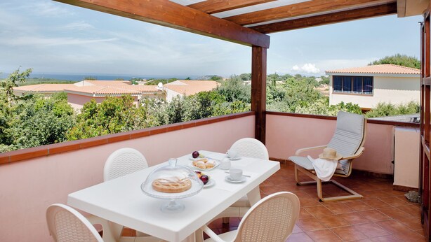 Un balcón con una mesa dispuesta para el desayuno, sillas blancas y vistas al mar y edificios circundantes.