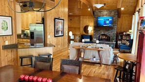 A cozy cabin interior with a kitchen, dining area, and living room, featuring wooden walls, a stone fireplace, and modern appliances. - Cashiers, NC Home on Golf Course w Trillium Club Access, Fast WiFi, Kid-Friendly (Cashiers)