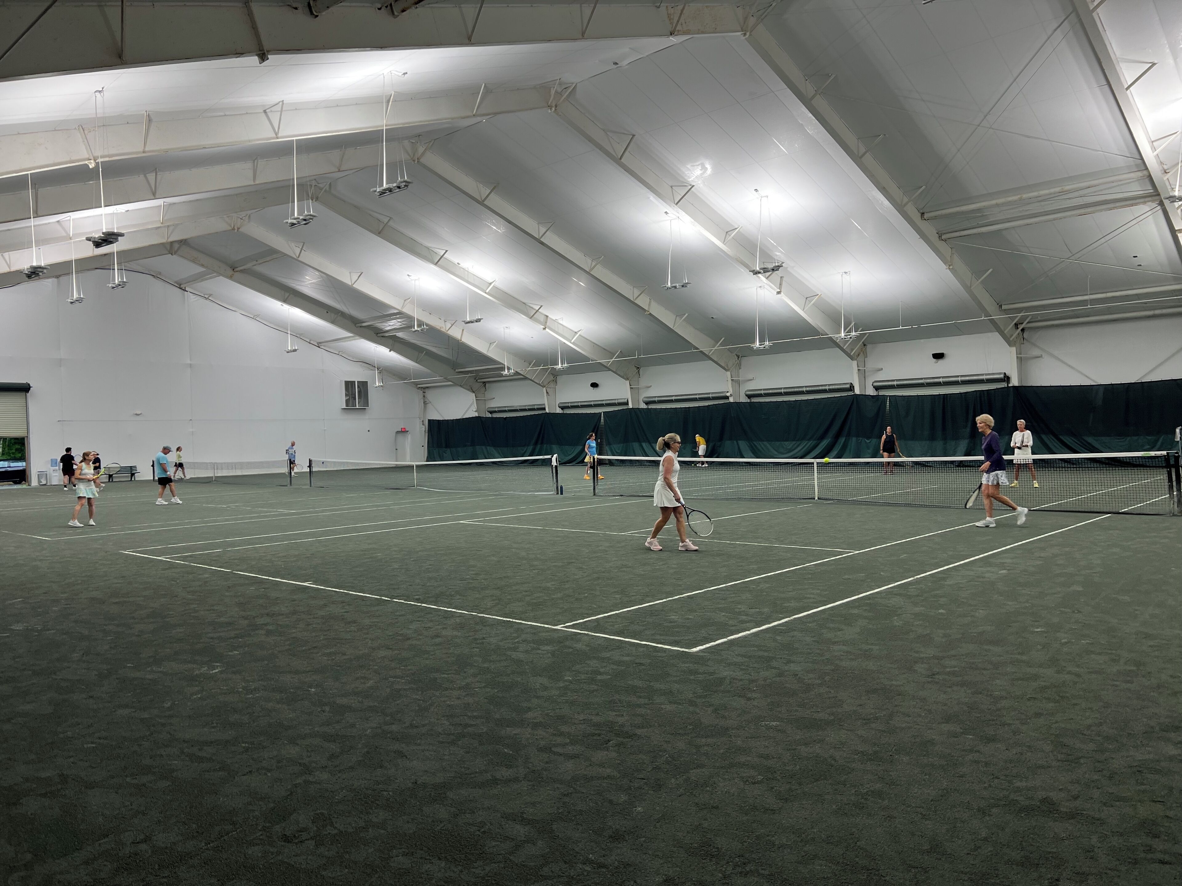 Una cancha de tenis interior con varios jugadores, una red e iluminación colgante.
