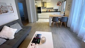 Un salon moderne avec un canapé gris, une table basse sur laquelle sont posés des livres et une bougie, et une cuisine en arrière-plan.