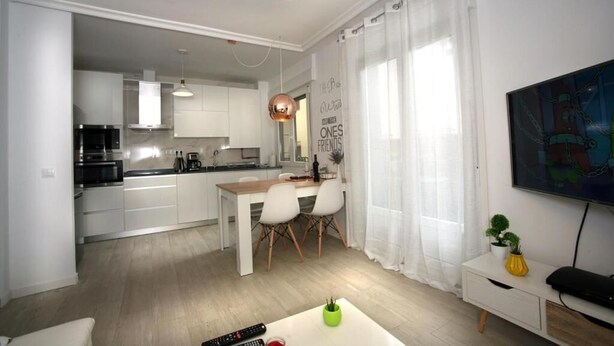 Une cuisine moderne avec des meubles blancs, un coin repas avec une table et des chaises en bois, et un salon avec un téléviseur et une plante.
