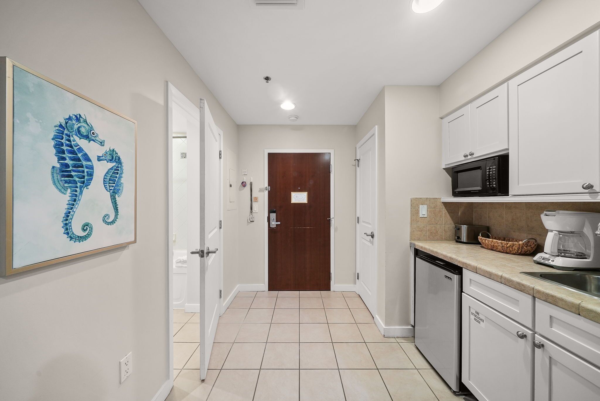 A narrow hallway with a wooden door at the end, white cabinets, a microwave, and a coffee maker on the counter.